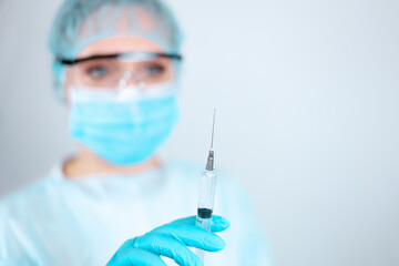 Fototapeta premium A nurse in a medical gown, mask and protective gloves with transparent glasses on her face looks at the needle of a syringe for injection
