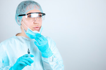 A nurse in a medical gown, mask and protective gloves with transparent glasses removes the mask from her face