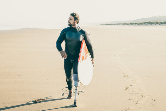 Active Surfer With Amputated Leg Walking On Beach With Surfboard. Bearded Amputee In Wetsuit Pacing On Sand, Carrying Board And Looking Away. Vertical Shot. Artificial Limb And Extreme Sport Concept