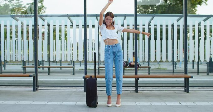 Medium shot of pretty woman with long hair standing outdoor with suitcase, headphones, listening music, dancing and smiling cheerfully at street. Portrait of beautiful happy stylish girl at bus stop.