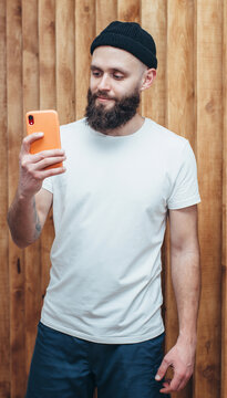 A young stylish guy, a hipster with a beautiful beard, dressed in a white, empty T-shirt with a place for your company or text, holds a smartphone and takes pictures of himself in the mirror