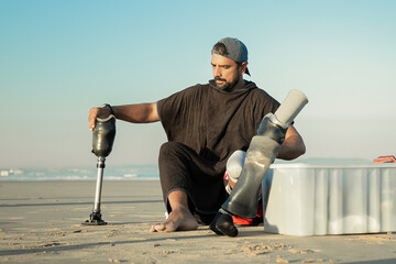 Serious male surfer sitting on sand near surfboard and changing below-knee prosthesis. Front view. Artificial limb and outdoor activity concept