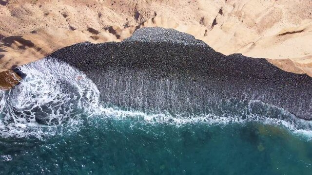 Vertical aerial shot view of a pebble beach and waves at the bottom of a cliff. Camera pulling back