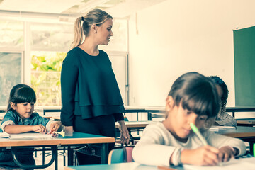 Serious female teacher watching primary schoolkids doing their task in class, sitting at desks and writing in copybooks. Education or back to school concept