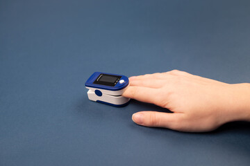 Hand with a finger in pulse oximeter on dark blue background. Self monitoring and protection during pandemic concept. Copy space.