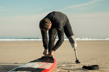Sporty man wearing wetsuit and artificial limb, tying board to his ankle on sand. Copy space. Artificial limb and active lifestyle concept