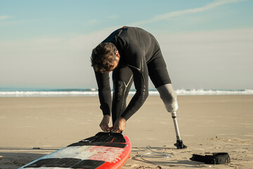 Sporty man wearing wetsuit and artificial limb, tying board to his ankle on sand. Copy space....