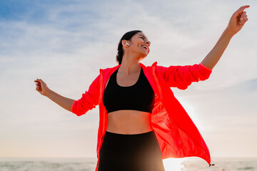 young smiling attractive slim woman doing sports in morning sunrise dancing on sea beach in sports...