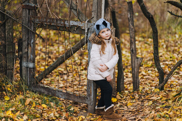A beautiful girl, a child in a white jacket and a hat stands and poses near a wooden gate in a village in nature in autumn against a background of yellow leaves. © shchus
