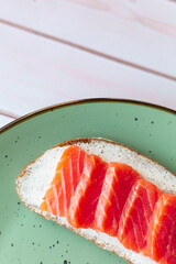 Toast with cream cheese, red fish salmon on a green ceramic plate on a white wooden background, breakfast