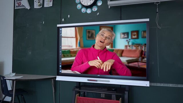 Female teacher gives a grammar lesson on an interactive whiteboard tv in classroom set against a blackboard in a school. Remote teaching learning concept.