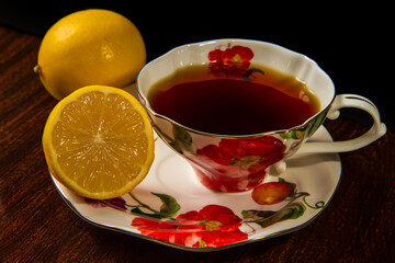 A porcelain cup with tea on a porcelain saucer. In the foreground is half a lemon. A whole lemon in the background.