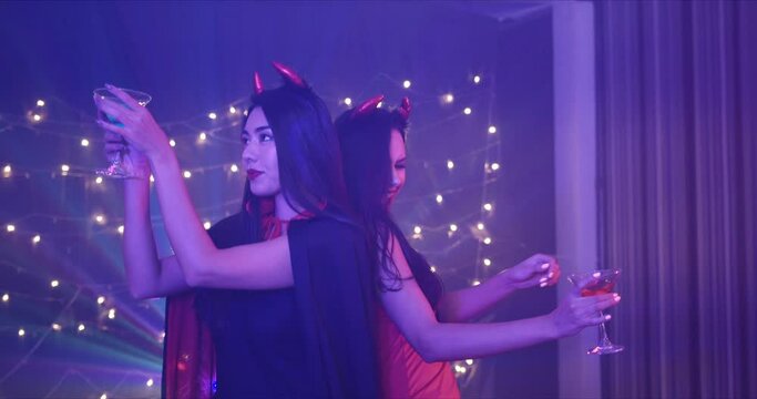 Medium Shot, Handheld - Two beautiful seductive young Caucasian female friends dressing in she devils seductively dancing at a Halloween costume party with drink in hand.