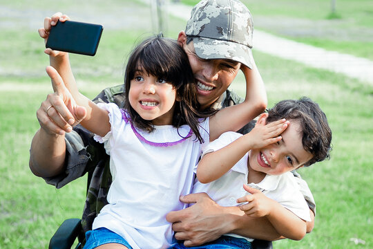 Two happy kids sitting on dads lap and taking selfie on cell. Disabled military man walking with kids in park. Veteran of war or disability concept