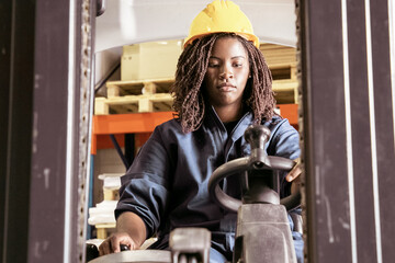 Focused young black female logistic worker in protective uniform operating fork lift in warehouse, pulling lever. Low angle. Female labor concept