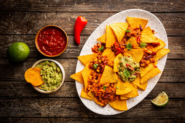 Delicious plate of yellow corn nachos chips with cheese, minced meat and red hot spicy salsa over wooden table, top view