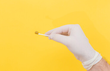 A doctor's hand in a white medical glove holds a stool sample on a stick on a yellow background. The medicine. Analysis. Cala. Infection. The close plan.