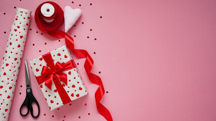 Valentine's day gift wrapping concept. Box wrapped in white paper in hearts,red bow, candy, red ribbon, reapers and a roll of wrapping paper on a pink background, top view, flat lay, copy space.