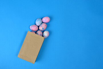 Easter eggs painted in blue and pink lie in a brown paper bag on a blue background, top view. Several colorful Easter eggs are spilled out of paper craft packaging