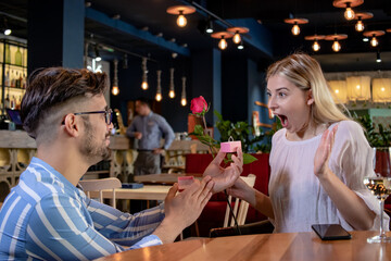 Young romantic couple on a date in a restaurant. Boyfriend making proposal to his girlfriend with a red rose and an engagement ring on valentine day.