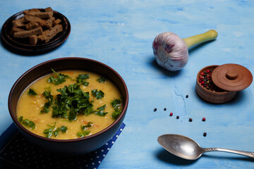 Bowl of pea or lentils soup served with fresh parsley, garlic, aromatic pepper set and rye bread crackers on blue wooden background. Healthy diet.