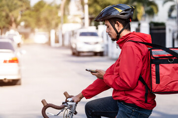 Obraz premium A courier in red uniform with delivery box on back riding bicycle and looking on cellphone to check addres to deliver food to customer. Courier on bicycle delivering food in city.
