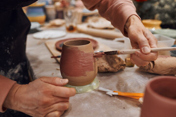 Fototapeta na wymiar Applying engobe on a ceramic mug with a brush in a pottery workshop. Hands close up