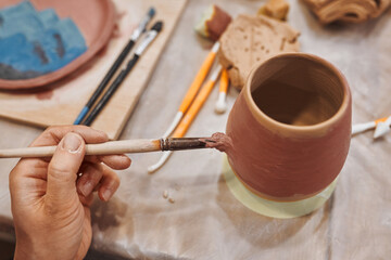 Applying engobe on a ceramic mug with a brush in a pottery workshop. Hands close up