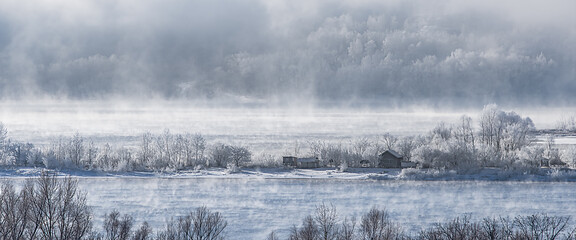 Winter frosty fog on an unfrozen river. Trees and houses in the snow on the shore.