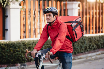 A courier in red uniform with delivery box on back riding bicycle and looking on cellphone to check...