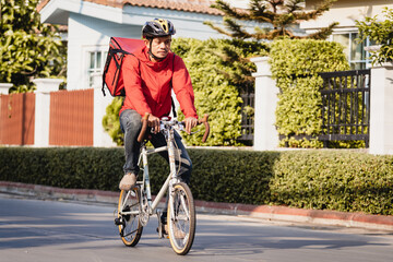 A courier in red uniform with delivery box on back riding bicycle and looking on cellphone to check addres to deliver food to customer. Courier on bicycle delivering food in city.