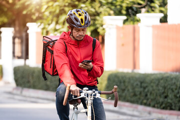 A courier in red uniform with delivery box on back riding bicycle and looking on cellphone to check addres to deliver food to customer. Courier on bicycle delivering food in city.