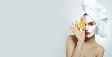 A beautiful young woman with a white towel on her head and a cosmetic cream mask on her face is holding a slice of lemon. Cosmetology.