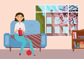 A young girl is engaged in knitting at home. Beautiful female character makes warm clothes with pleasure while sitting in a chair by the window. Person is engaged in a hobby during her free time