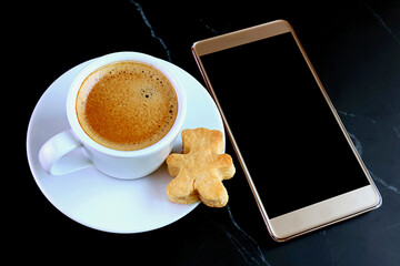 Cup of Hot Coffee and Teddy Bear Shaped Cookie with Blank Screen Mobile Phone on Black Table	