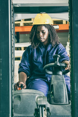 Focused young black female logistic worker in hardhat operating fork lift in warehouse, pulling lever. Front view. Female labor concept