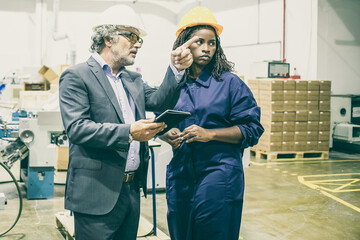Content senior boss standing, talking with female plant worker and holding tablet. Successful pensive bearded man explaining task to African American employee. Manufacture and occupation concept