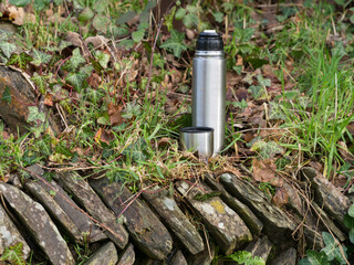 A silver metal flask and cup stand on a stone wall in a rural setting