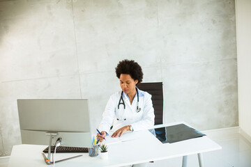 Female African American doctor wearing white coat with stethoscope sitting behind desk in office