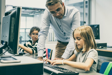 Obraz premium Focused pretty blonde girl typing on keyboard in classroom. Serious middle-aged professor in glasses looking at her, standing near desk and explaining task. Informatics and education concept