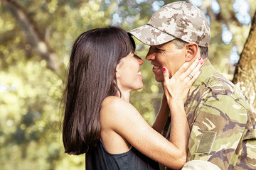 Happy military man and his wife hugging and kissing in city park. Side view, medium shot. Returning home or relationship concept