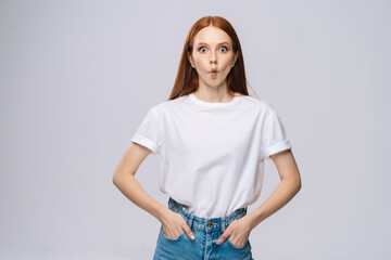 Obraz premium Beautiful young woman wearing T-shirt and denim pants looking at camera with big eyes and fish lip on isolated white background. Pretty lady model with red hair emotionally showing facial expressions.