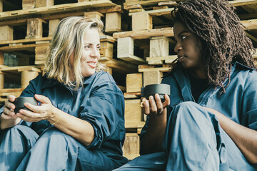 Positive female factory workers chatting while drinking coffee, sitting at wooden platforms in warehouse. Front view. Labor or coffee break concept