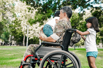 Fototapeta na wymiar Middle aged disabled military dad walking with two children in park. Girl holding wheelchair handles, boy standing on dads lap. Veteran of war or disability concept