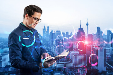 Eastern young businessman looking into his notebook to achieve tremendous growth in business based on technologies. Worldwide process to conduct transactions. Tech hologram icons over Kuala Lumpur