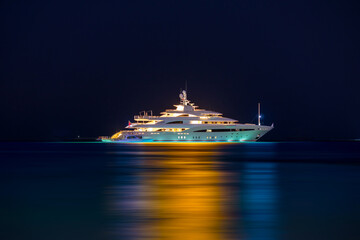 Night view to large illuminated white boat located over horizon, colorful lights coming from yacht reflect on the surface of the the Gulf sea. Shot at blue hour.  