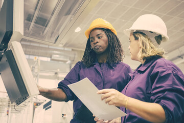 Black female industrial employee teaching colleague to operate machine, pointing at monitors over...
