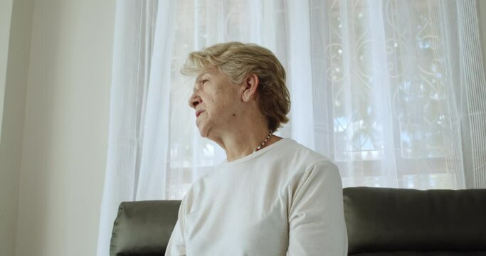 An elderly woman after retirement cut short hair with light brown hair wearing a round neck long a sleeve T-shirt and a necklace, sit on a brown sofa pain, sore muscles in the shoulder blades at home.