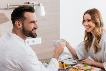 Obraz na płótnie Canvas Smiling man in bathrobe holding cup near breakfast and girlfriend on blurred background in hotel
