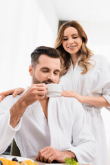 Man holding cup of coffee near smiling girlfriend in bathrobe on blurred background in hotel